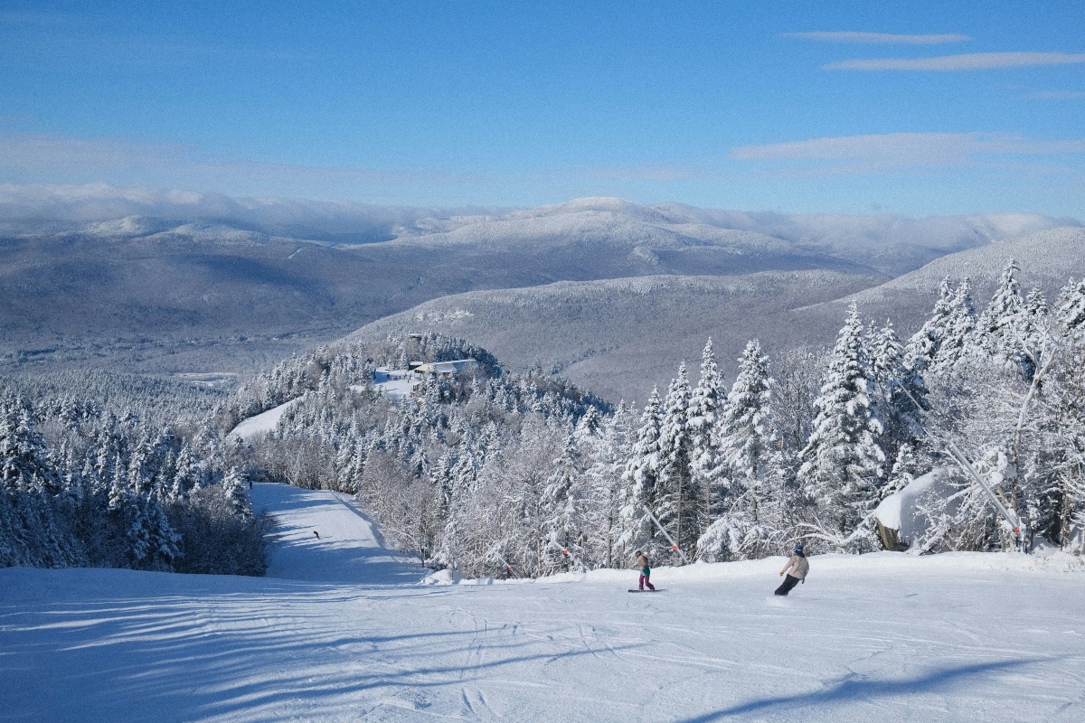 Two snowboarders make their way down a groomed ski trail lined with snow-covered evergreens on a clear winter day. Beyond the slope, a small lodge sits on a ridge and rows of white mountain ridgelines recede into the distance under a bright blue sky.