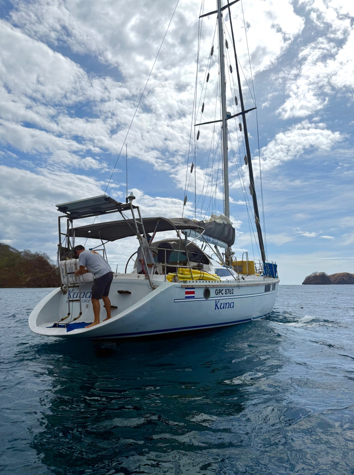 A white sailing catamaran named Kuna sits on calm blue-green water, with a crew member in a gray shirt working at the stern platform. Rocky headlands are visible on both sides of the horizon under a partly cloudy sky.