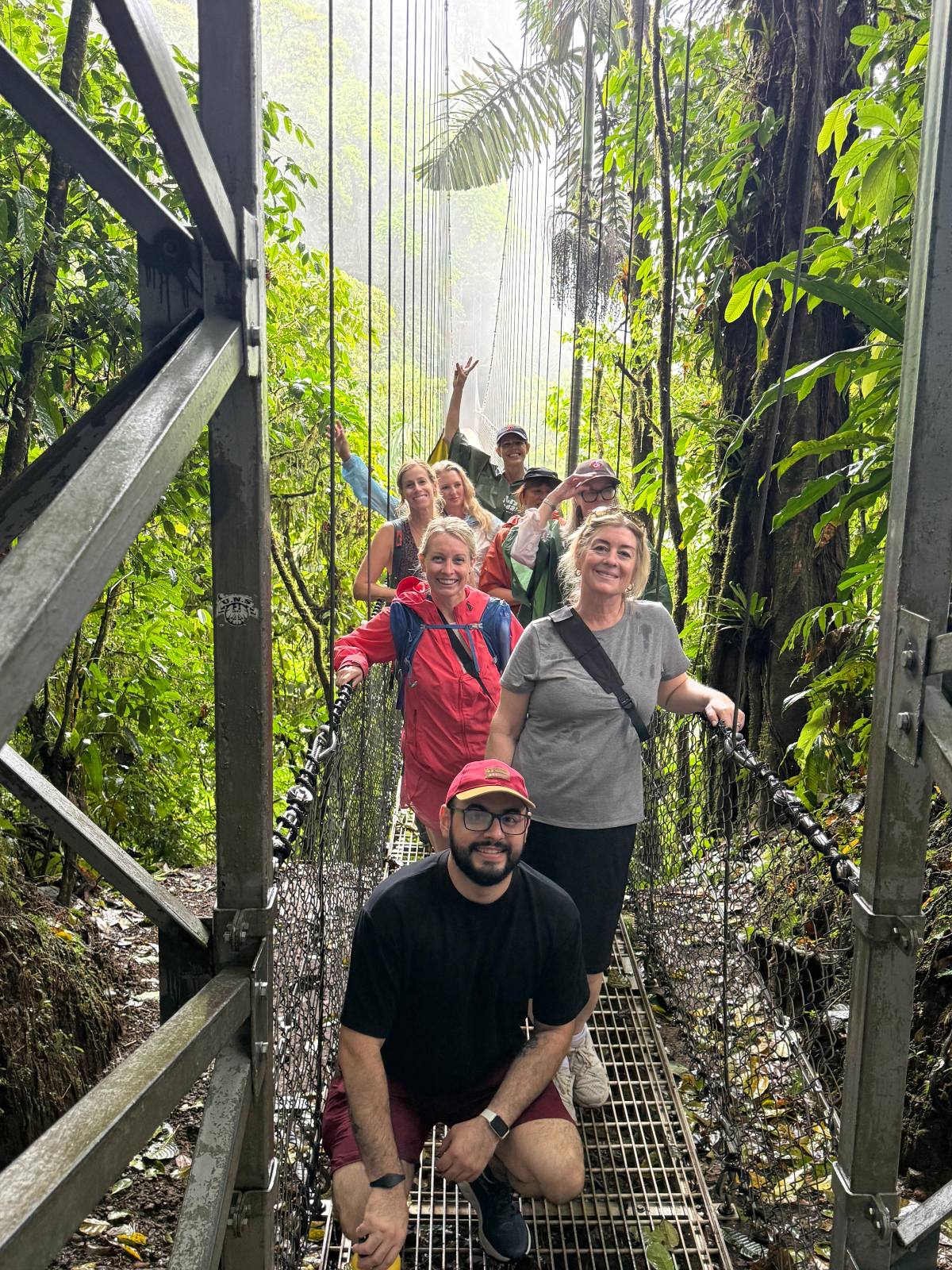 A group of about nine people poses on a metal suspension bridge in the rainforest, several smiling toward the camera while one raises an arm overhead. A man in a red cap crouches at the front of the group, with dense green foliage, hanging vines, and mist filling the background.