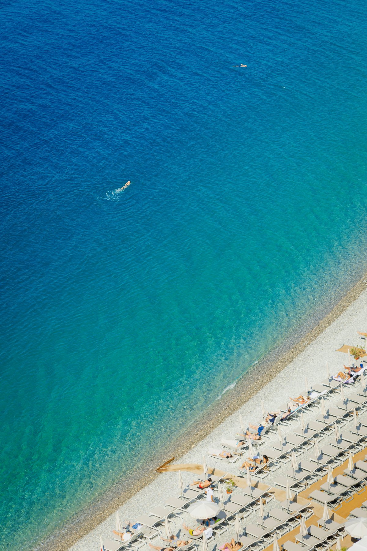 Aerial view of clear Mediterranean water shifting from deep blue to pale turquoise as it meets a narrow pebble beach, with two swimmers visible offshore. Along the shoreline, rows of white lounge chairs and folded umbrellas are arranged in angled lines, with a handful of sunbathers scattered among them.