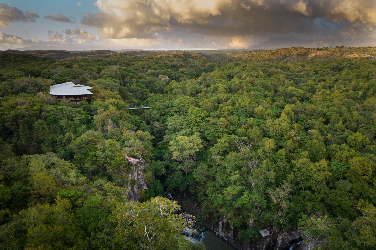 An aerial view of a forested river canyon in Costa Rica, with a lodge building topped by a wide white roof set on the ridge at left and a suspension footbridge spanning the gorge beside it. A zipline cable crosses the canyon above the river below, with dense green tree canopy stretching to distant hills and mountains under a cloud-filled sky at dusk.