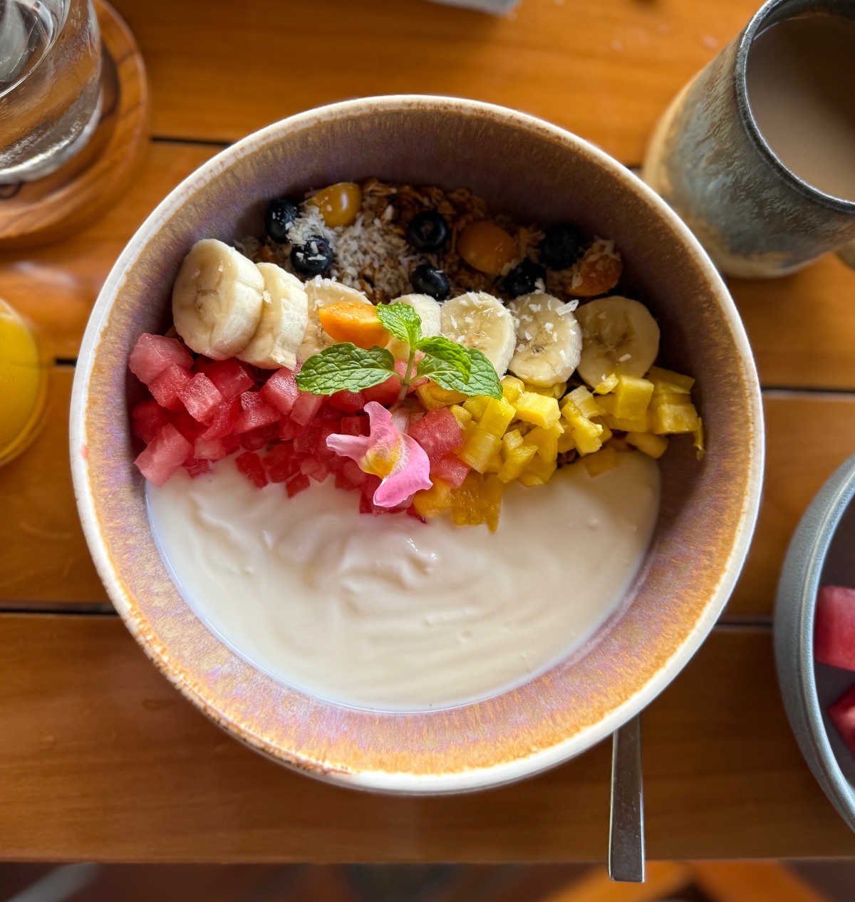 An overhead view of a ceramic bowl of yogurt topped with sliced banana, diced watermelon and pineapple, blueberries, granola, shredded coconut, a sprig of mint, and a pink edible flower. The bowl sits on a wooden table alongside a spoon, a glass of juice, a stoneware mug of coffee, and a second bowl of cut fruit at the edge of the frame.