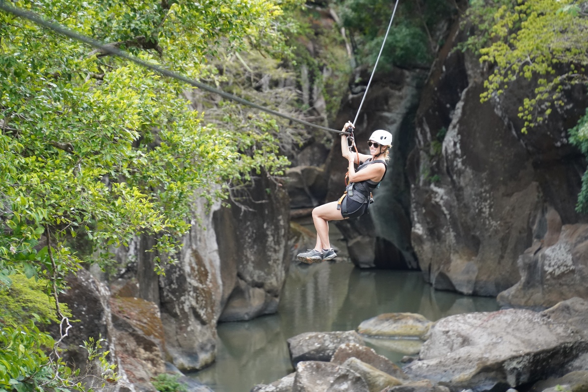 A woman in a white helmet, sunglasses, and a harness rides a zipline cable across a narrow river canyon, holding the line overhead with her legs tucked beneath her. Sheer gray rock walls rise on either side above a shallow green river and scattered boulders, with leafy branches framing the left side of the frame.