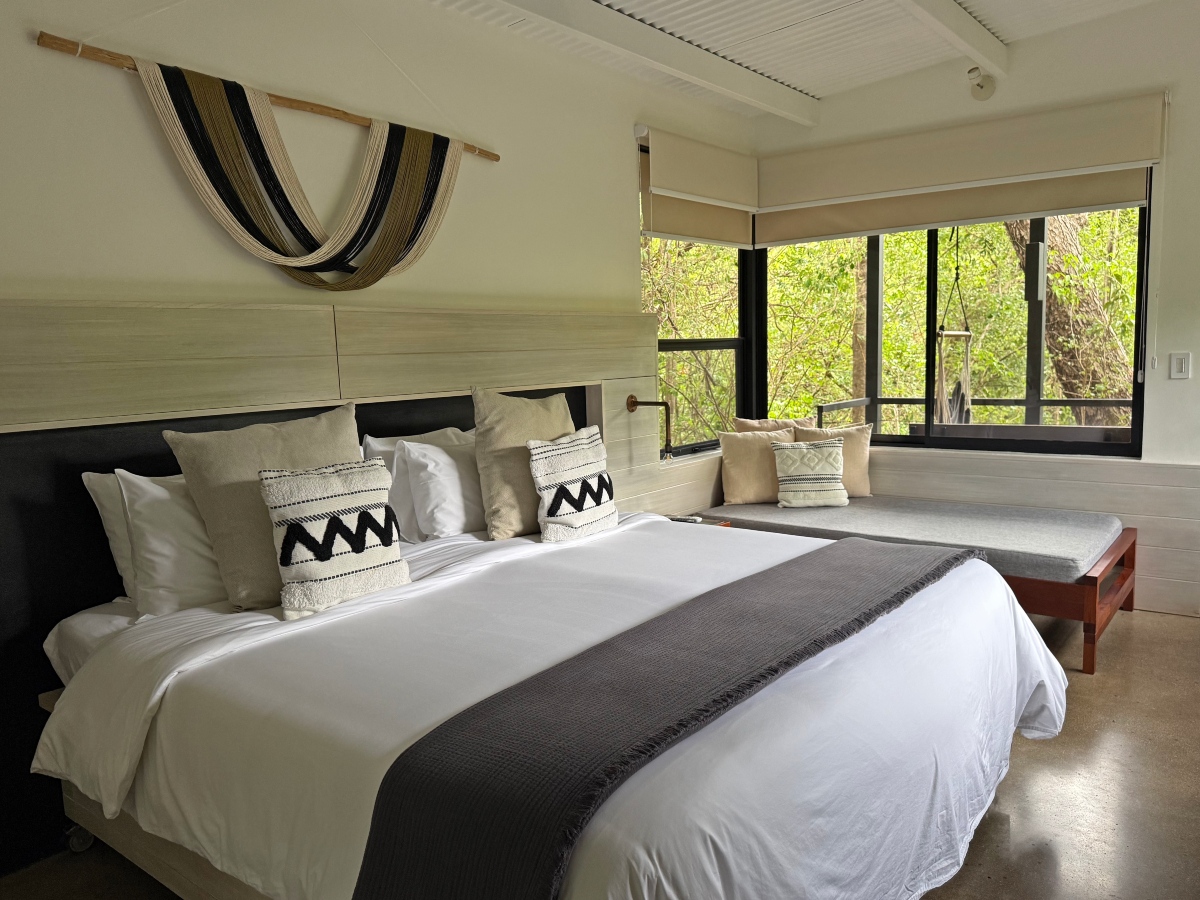 A guest room with a king bed made up in white linens, topped with a dark gray fringed throw and layered beige and black-and-white patterned pillows against a light wood headboard panel. A striped woven hammock hangs as wall decor above the bed, and a cushioned bench sits beneath a large window looking out onto green forest, with a polished concrete floor and a white slatted ceiling.