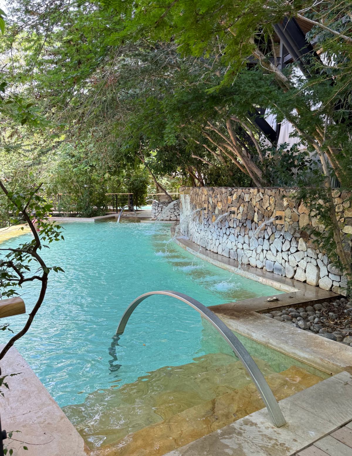 A curved outdoor pool with turquoise water and a shallow stepped entry with a metal handrail in the foreground. A stacked stone retaining wall runs along the right edge with several spouts pouring water into the pool, and leafy trees shade the deck and surrounding area.