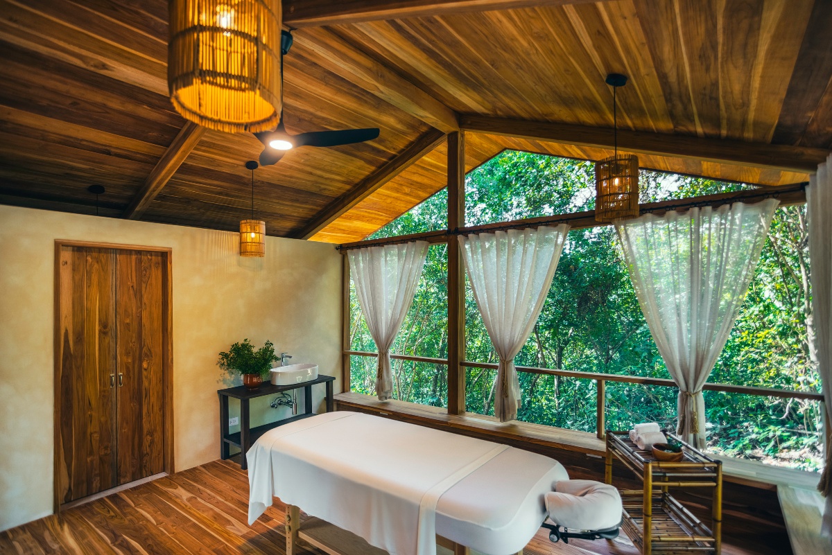 A spa treatment room with a massage table draped in white linens beneath a peaked wood ceiling hung with woven pendant lights and a ceiling fan. Open windows with sheer tied-back curtains line the far wall and look out into green forest, with a wooden door, a small washbasin console with a potted plant, and a bamboo cart of towels along the sides.