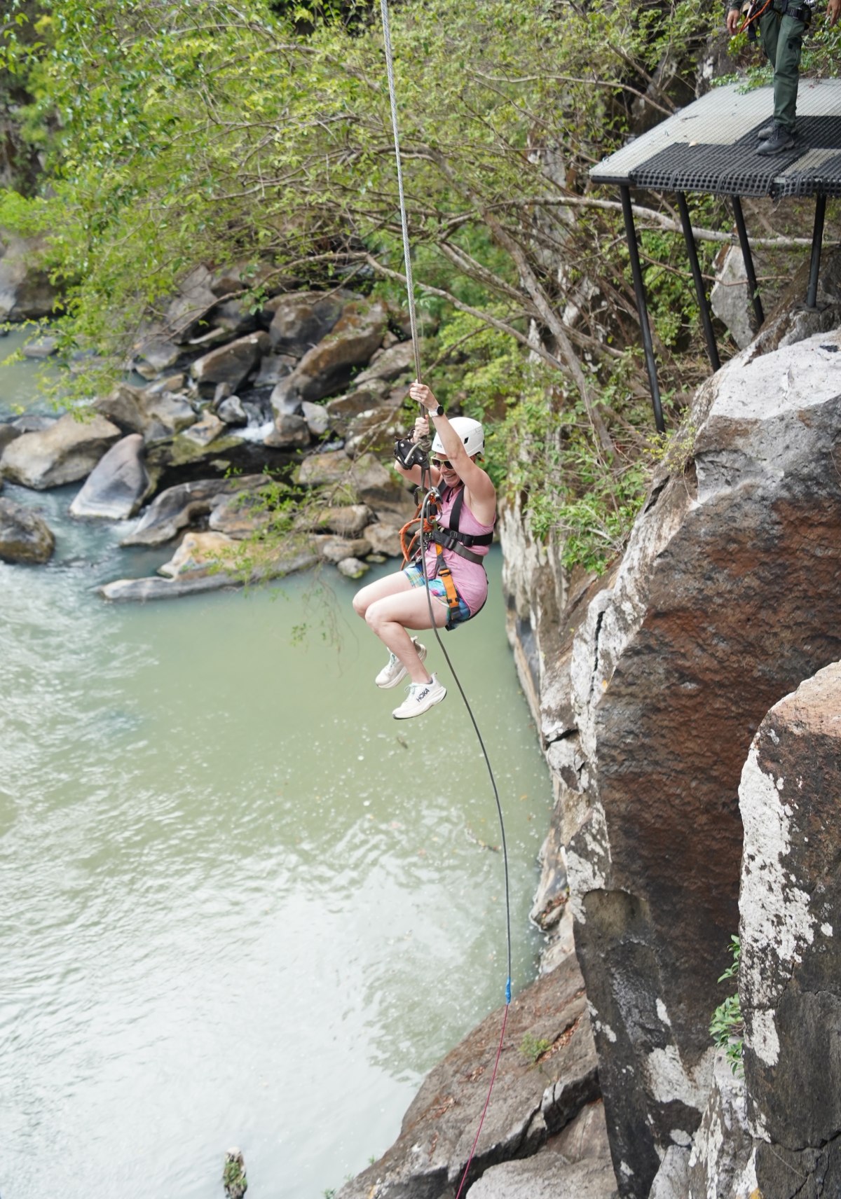 A woman in a helmet, harness, pink tank top, and shorts swings on a cable high above a river canyon, gripping the line with her knees bent. A guide stands on a small metal platform cantilevered from the rock face above her, with pale green water, boulders, and green trees filling the canyon below.