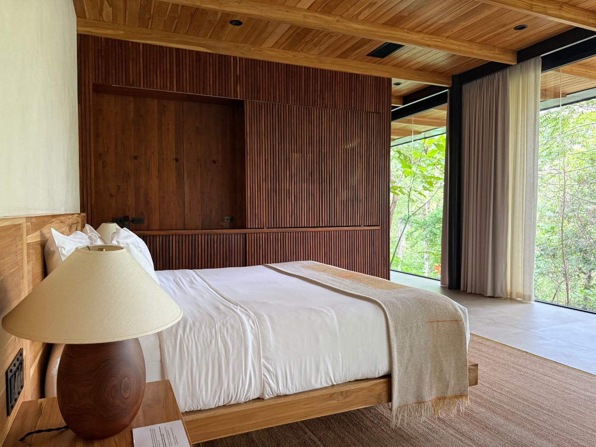 A guest room with a low wooden platform bed dressed in white linens and a fringed cream throw, set against a dark slatted wood wall beneath a plank ceiling with exposed beams. A rounded wood table lamp sits on a nightstand in the foreground, and floor-to-ceiling glass with sheer and solid curtains opens onto green forest along the far wall.