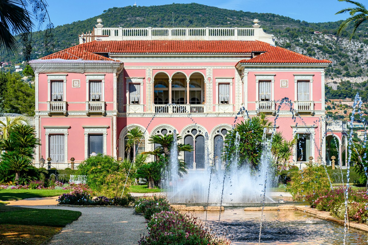 A pink Belle Époque villa with white stone trim, arched windows, and a terracotta tile roof stands behind a formal garden, with a wooded hillside rising in the background. In the foreground, jets of water arc from a long reflecting pool bordered by flower beds, palms, and a gravel path.