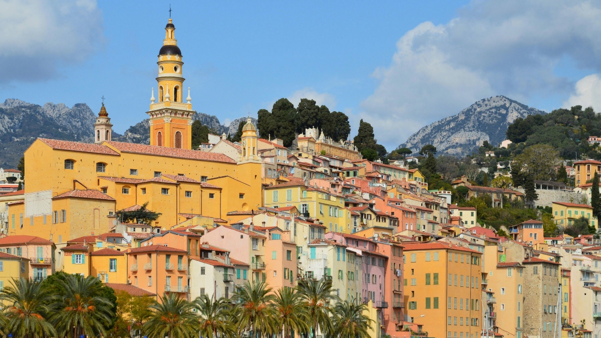 Menton's old town rises in tiers of pastel yellow, pink, and peach buildings with terracotta roofs and shuttered windows, topped by a large ochre church with a tall baroque bell tower. Palm trees line the base of the hillside, and rocky gray mountains rise behind the town under a partly cloudy sky.