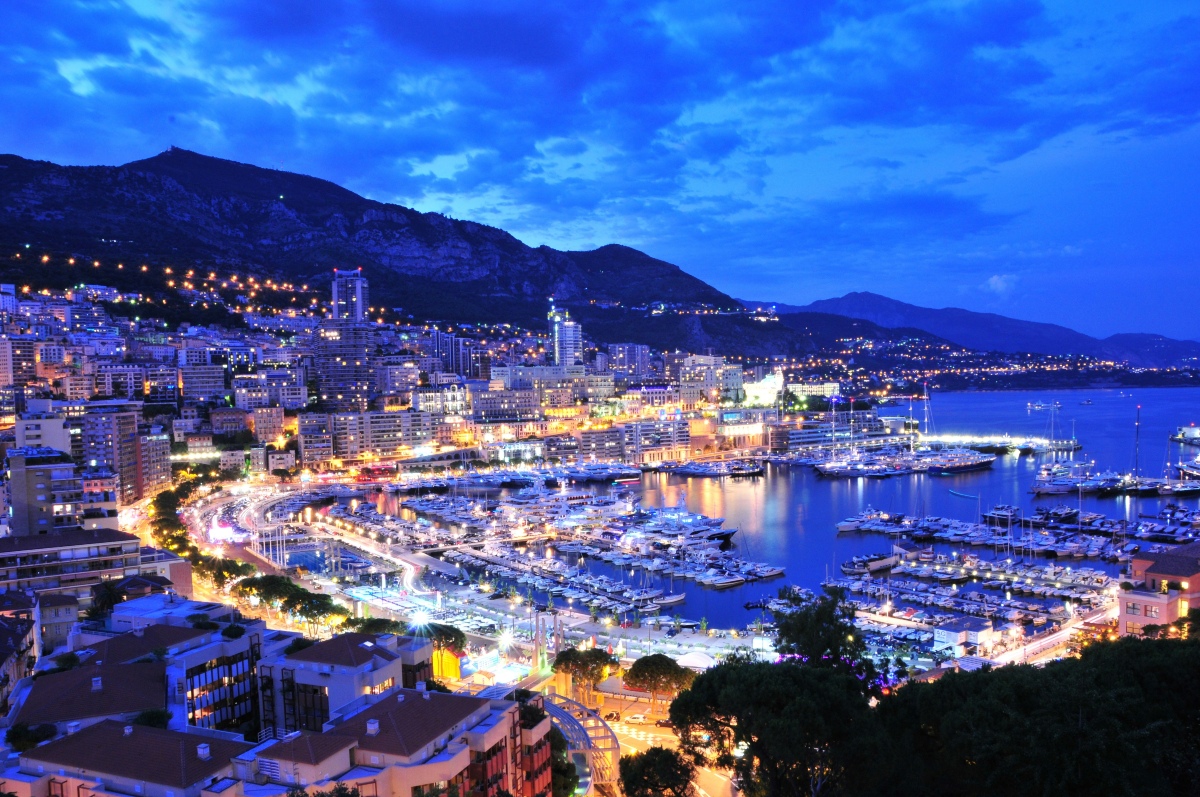 Elevated view of Monaco at dusk, with high-rise buildings and hillside roads lit in gold against a deep blue sky and dark mountains. Below, yachts and boats fill the harbor, their lights reflecting on the still water.