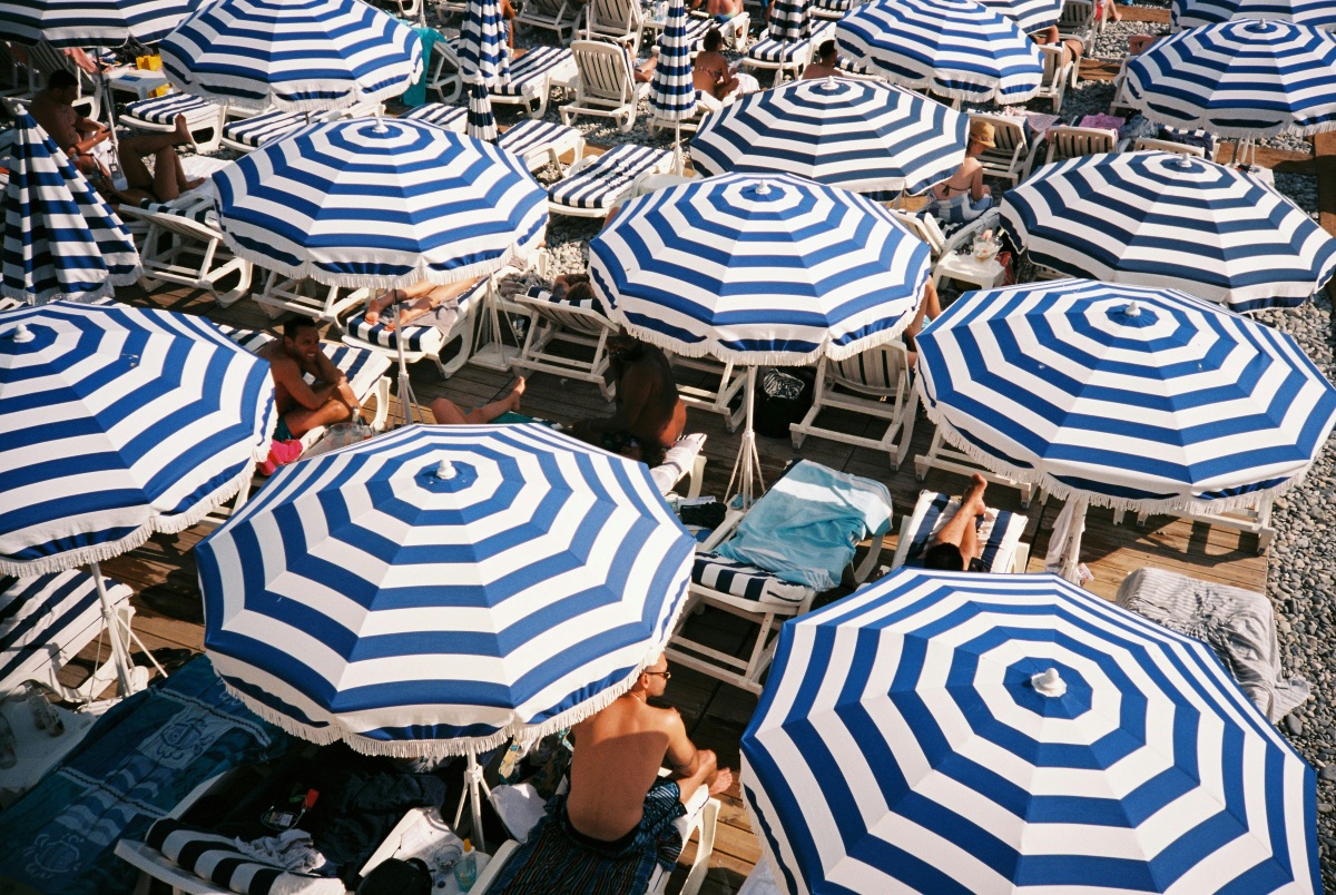Overhead view of a beach club densely packed with blue-and-white striped umbrellas, their fringed canopies overlapping across wooden decking and pebbles. Beneath and between them, sunbathers sit and recline on white loungers with towels, bags, and beach gear scattered nearby.