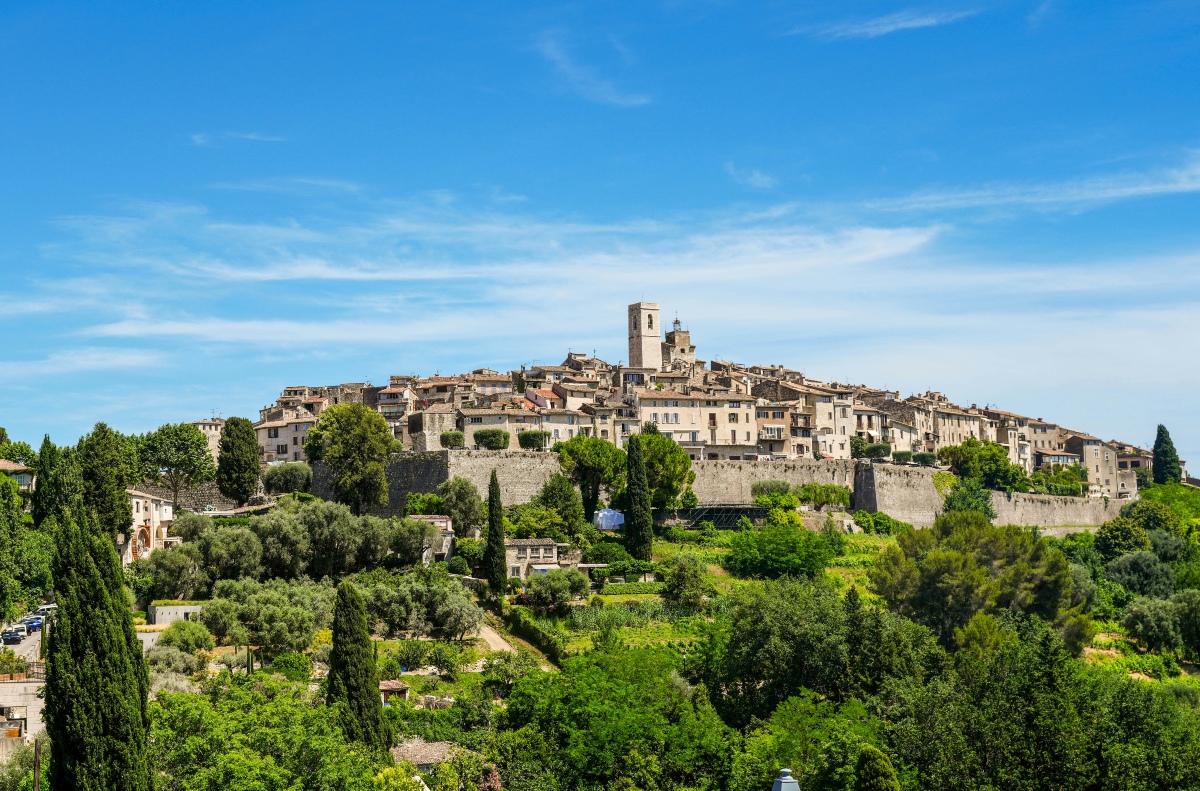 The hilltop village of Saint-Paul-de-Vence sits atop a ridge, its tightly packed stone buildings and terracotta roofs enclosed by medieval ramparts, with a square bell tower rising at the center. Below the walls, cypress trees, olive groves, and terraced greenery cover the slope under a clear blue sky.
