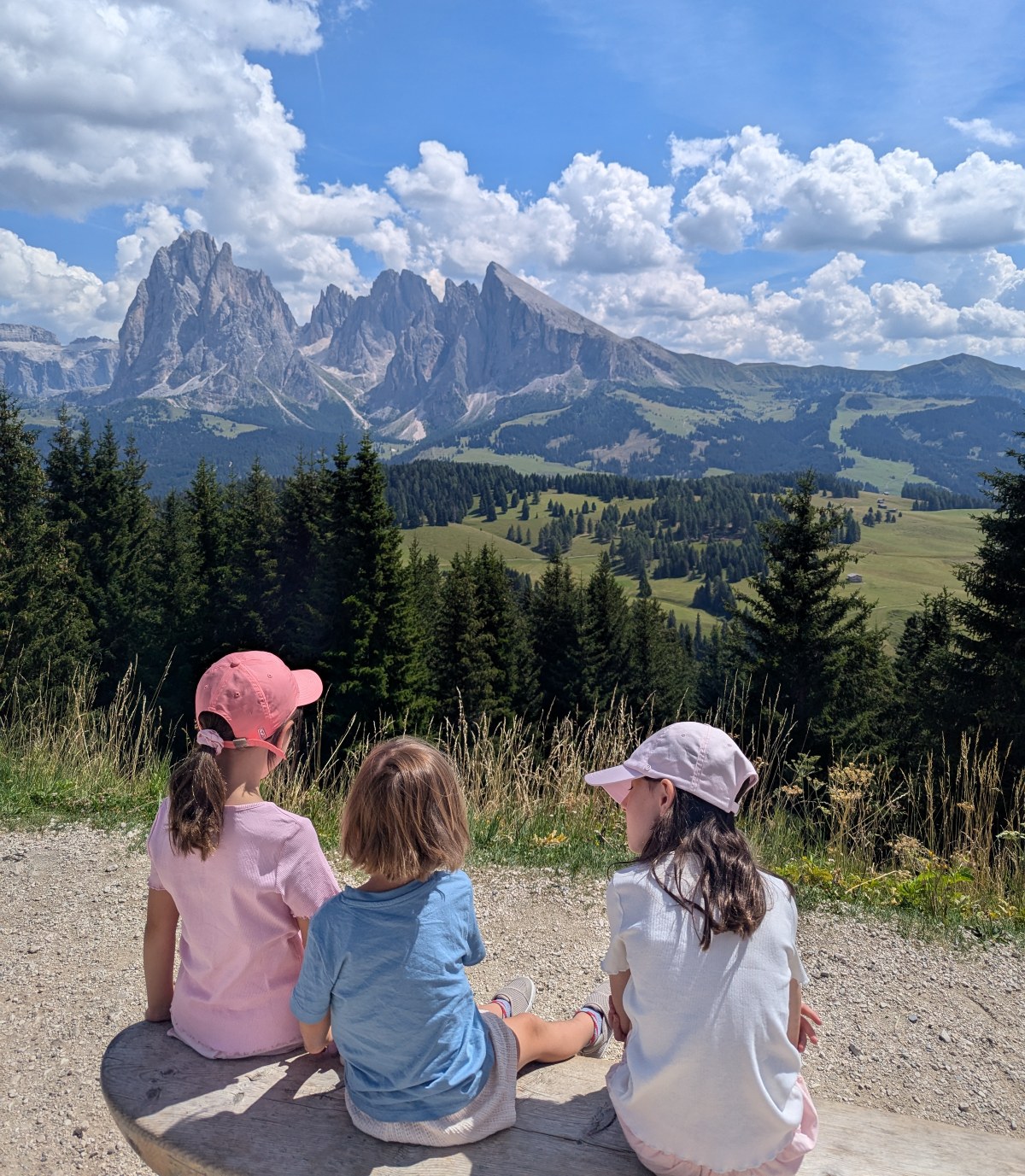 Three children sit side by side on a wooden bench, seen from behind, looking out over a Dolomites landscape of jagged limestone peaks, rolling green alpine meadows, and dense evergreen forest. Two of the children wear pink baseball caps and the child in the middle wears a blue T-shirt, all beneath a bright blue sky scattered with puffy white clouds.
