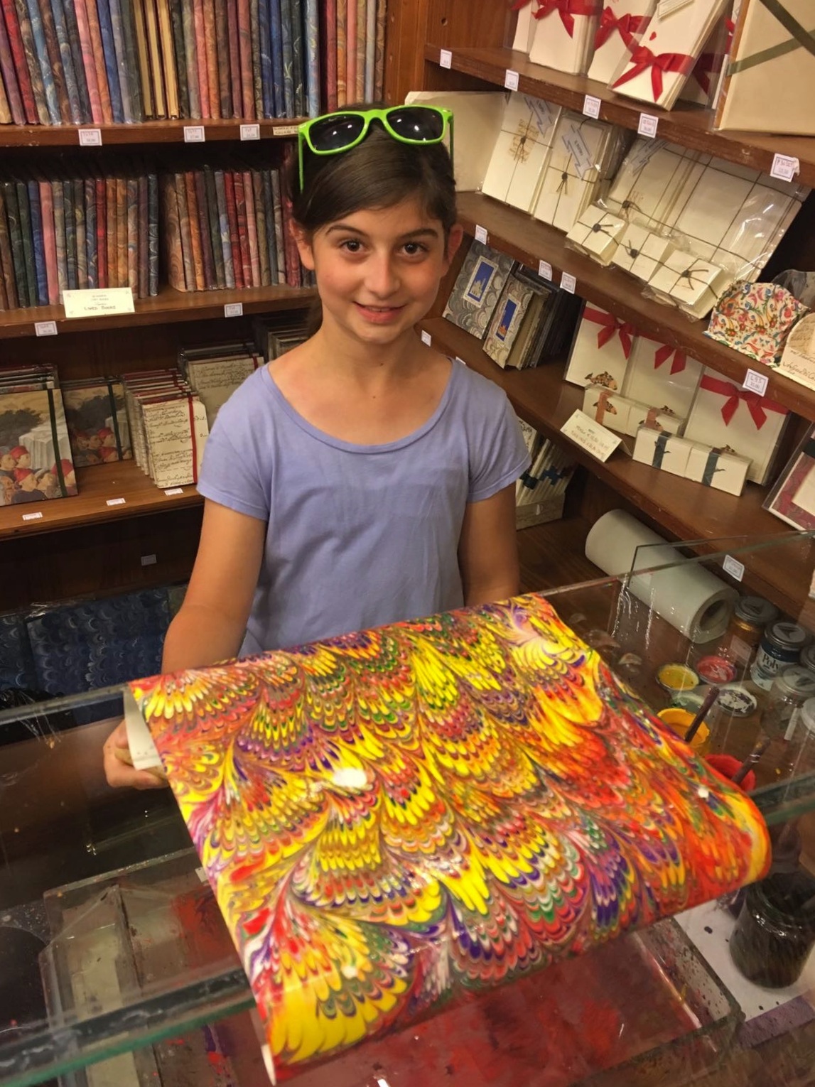 A girl in a lavender T-shirt with green sunglasses pushed onto her head holds up a freshly made sheet of marbled paper in vivid yellow, red, purple, and green feather patterns. She stands in a paper shop behind a shallow marbling tray, with wooden shelves of patterned notebooks, stationery sets, and ribbon-tied boxes filling the background.