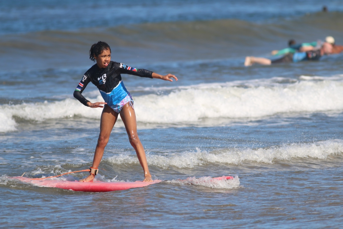 A girl in a black and blue long-sleeve rash guard stands balanced on a pink foam surfboard with arms outstretched, riding a small whitewater wave close to shore. An ankle leash connects her to the board, and another surfer lies paddling on a board in the background.