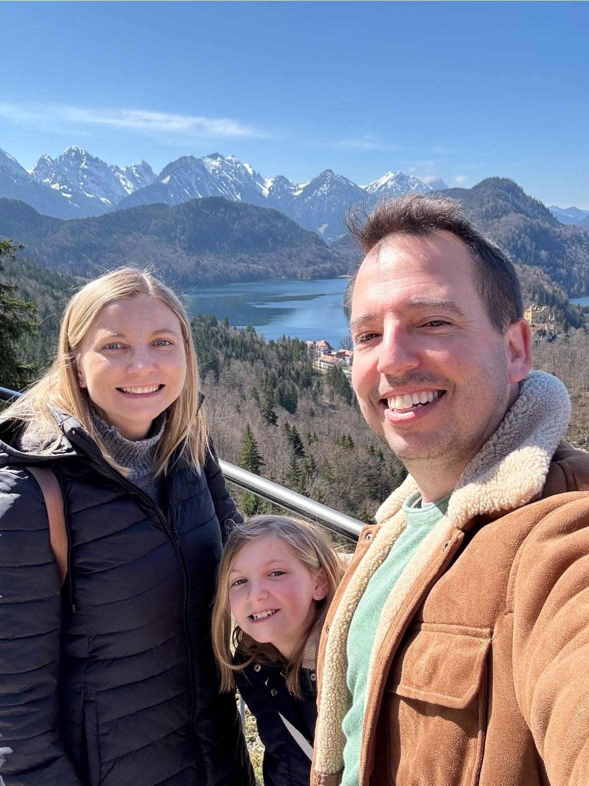 A family of three takes a selfie at a railed overlook, with a woman in a black puffer jacket, a girl with long blonde hair between them, and a man in a tan sherpa-lined jacket closest to the camera. Behind them stretch snow-capped Alpine peaks, evergreen-covered hillsides, and a deep blue lake under a clear sky.