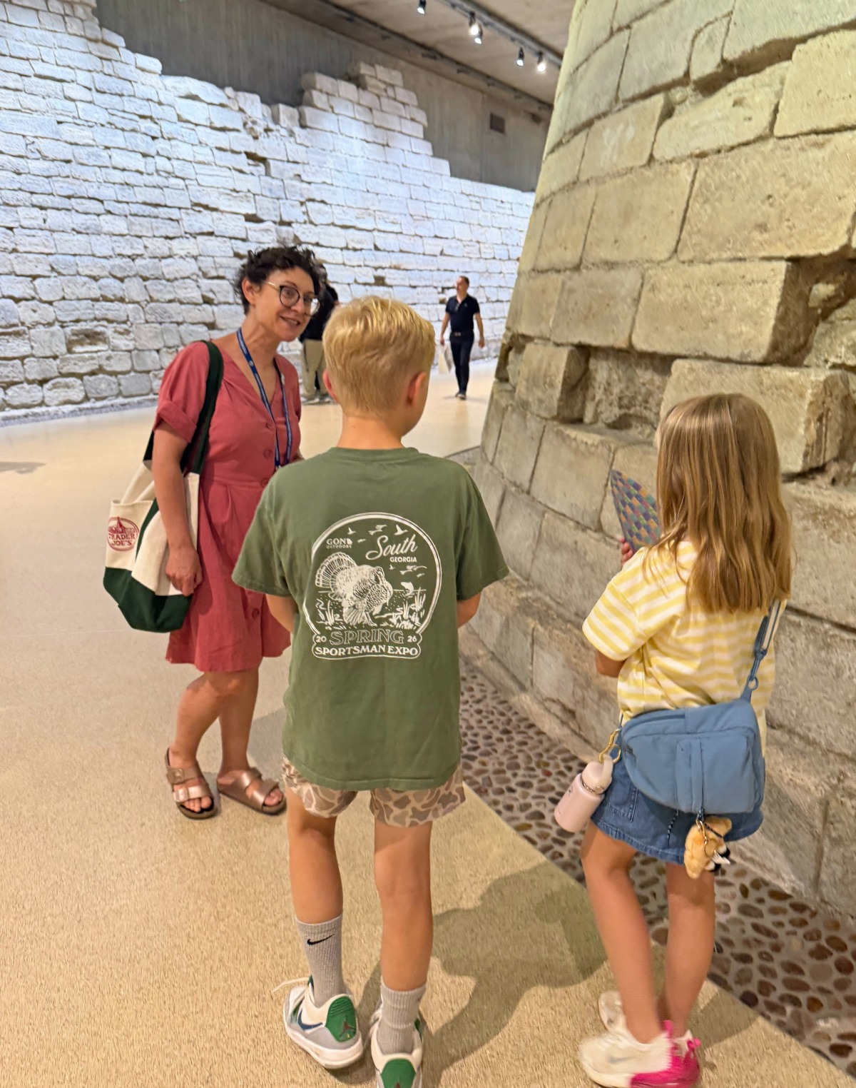 A woman in a rose-colored dress with a lanyard and tote bag speaks with two children inside an indoor archaeological site, where excavated ancient stone walls rise behind them. The boy, seen from behind, wears a green graphic T-shirt and camo shorts, while the girl in a yellow striped shirt and denim skirt carries a light blue crossbody bag and looks toward the stonework.