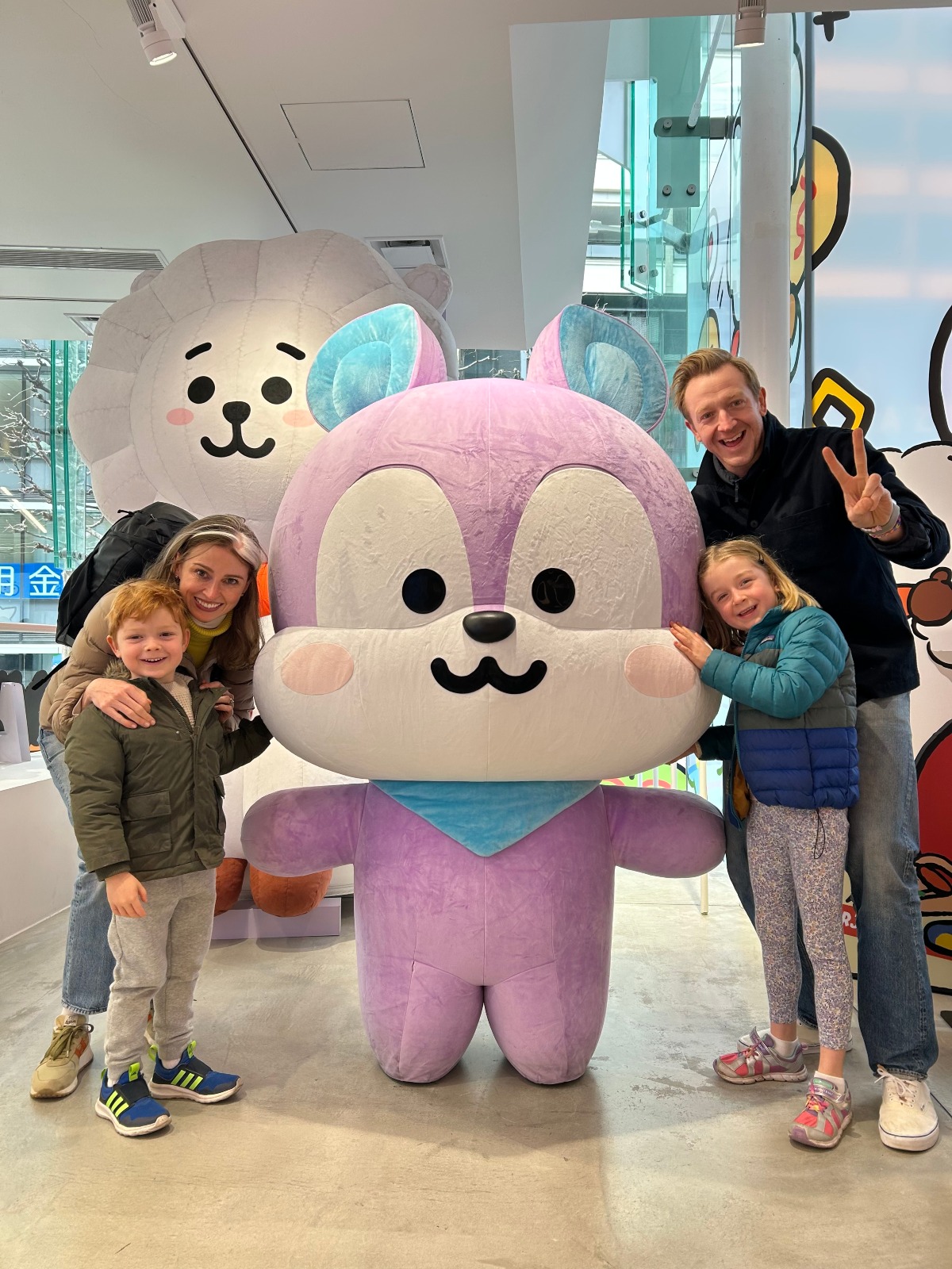 A family of four poses on either side of a large purple and white plush character statue inside a bright character-themed store, with a woman and young boy in a green jacket on the left and a man flashing a peace sign beside a girl in a teal and blue puffer vest on the right. A second oversized cream-colored plush figure and colorful cartoon graphics on the windows fill the background.
