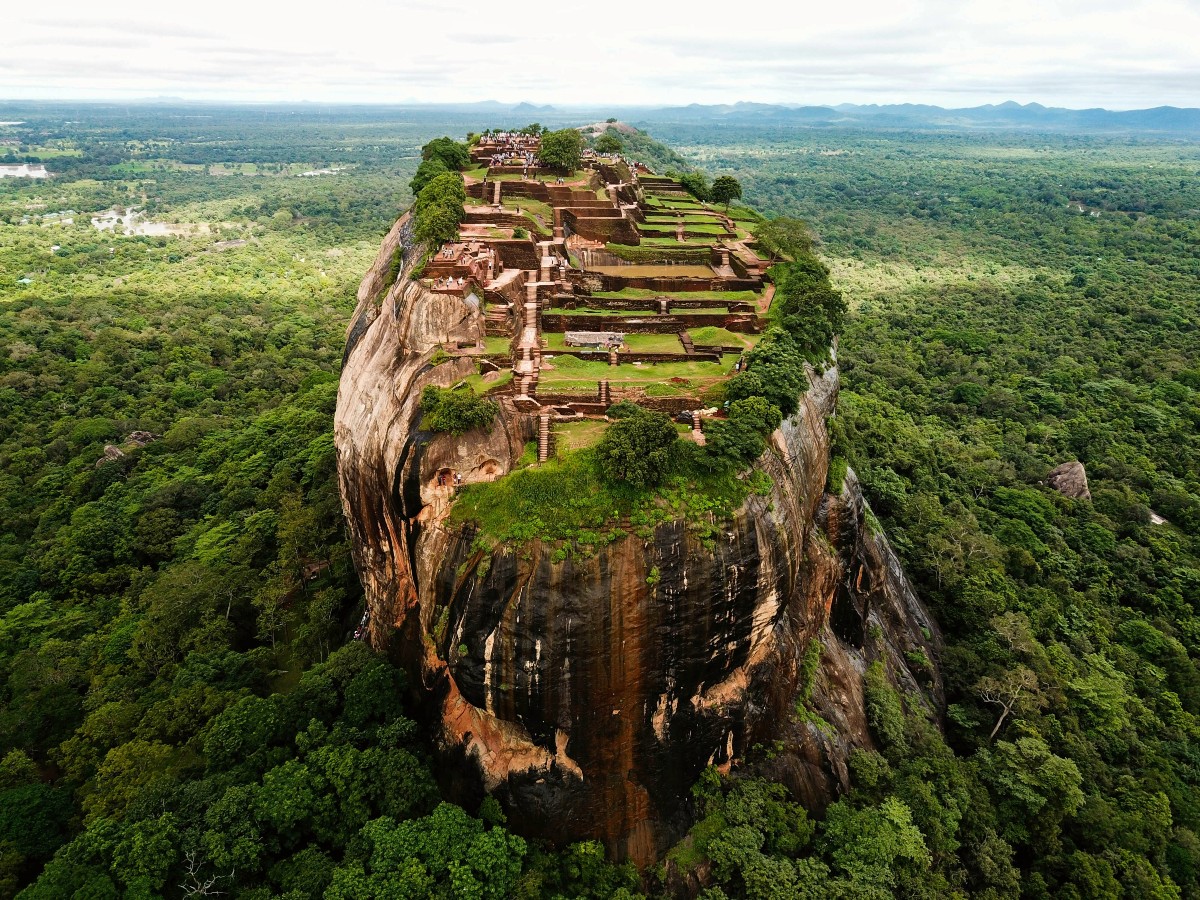 An aerial view of Sigiriya Rock, a sheer-sided granite outcrop rising abruptly from flat green jungle, with the terraced brick ruins of a palace complex covering its flat summit. Stone staircases and foundation walls step down the length of the plateau, and jungle stretches to distant hills on the horizon.