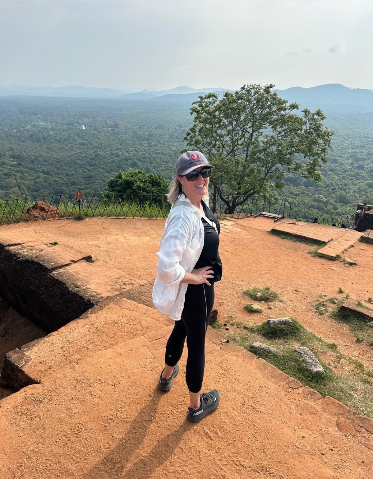 A woman in a white linen shirt, black tank top, black leggings, sunglasses, and a gray Ciao Bambino baseball cap stands on the flat orange-brick summit of Sigiriya Rock, turning back toward the camera with one hand on her hip. Behind her, a low iron railing edges the plateau and a lone flat-topped tree rises above dense green jungle that stretches to hazy blue mountains on the horizon.
