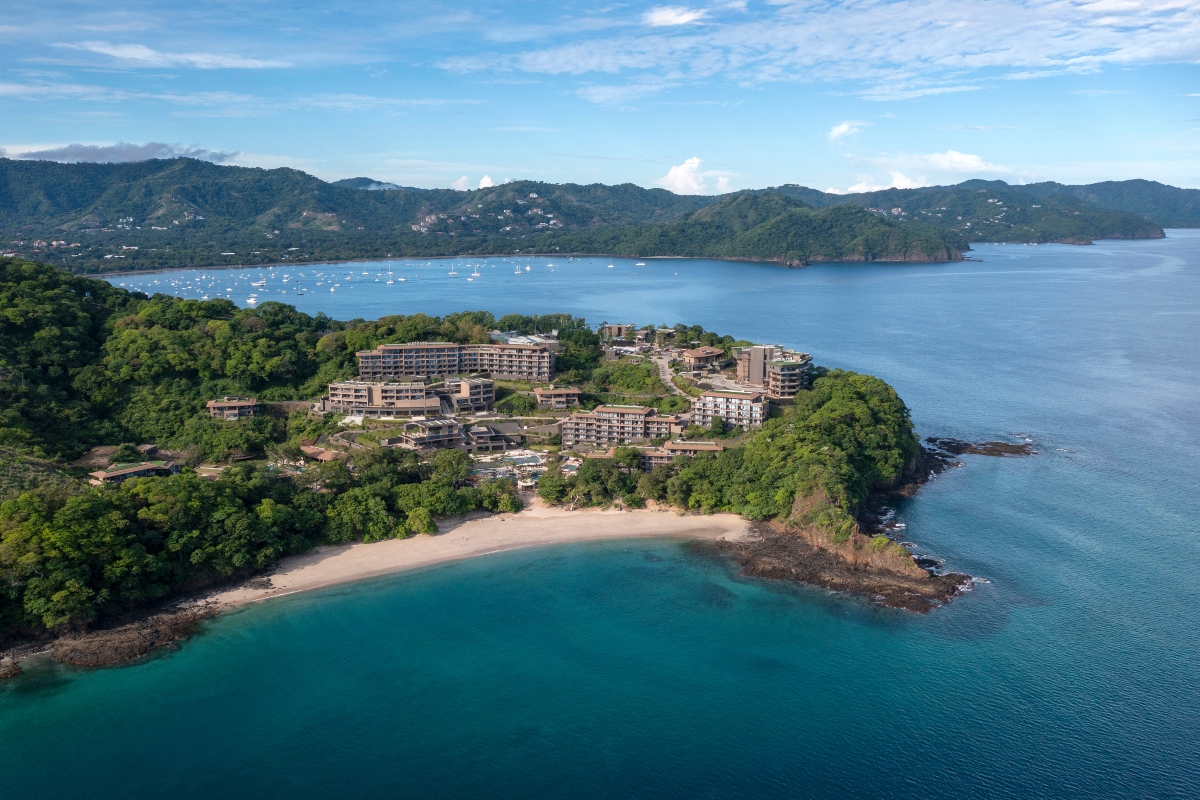 An aerial view of a forested peninsula holding a low-rise resort of tiered stone-and-wood buildings, set among dense green treetops above a crescent of pale sand beach. Turquoise water curves around the rocky point in the foreground, while a wide bay dotted with anchored sailboats stretches toward green coastal hills under a partly cloudy sky.