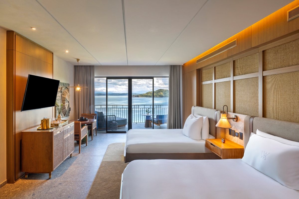 A hotel guest room with two beds made in white linens, monogrammed pillows, and upholstered headboards set against a wall of woven cane panels lit by warm cove lighting. Across the room, a wood cabinet holds a wall-mounted television beside a small desk, and floor-to-ceiling glass doors open onto a balcony with seating and a view of the ocean and a green headland.