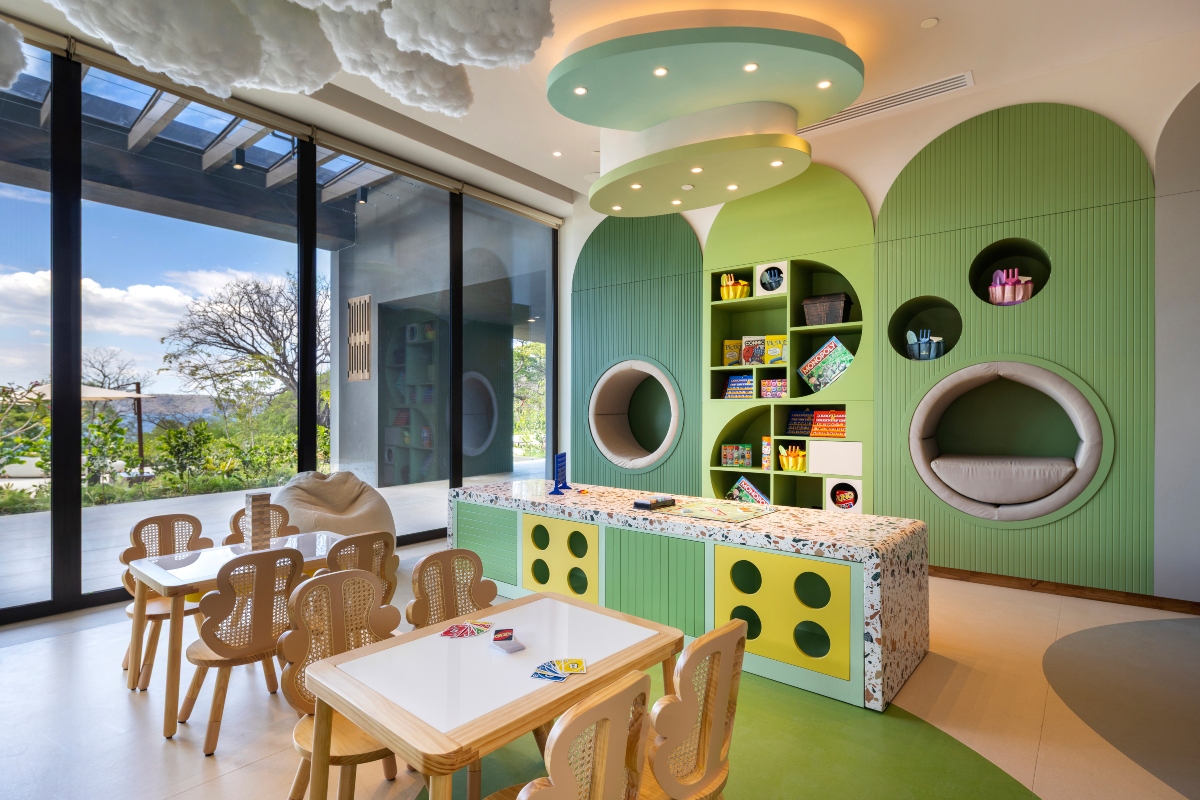 A children's playroom with two low tables and cane-backed kid-sized chairs, a terrazzo-topped counter, and a green arched wall built with cubby shelves of board games and craft supplies plus two padded circular nooks. Cloud-shaped sculptures hang from the ceiling above floor-to-ceiling windows that look out to trees and a distant view of the water.