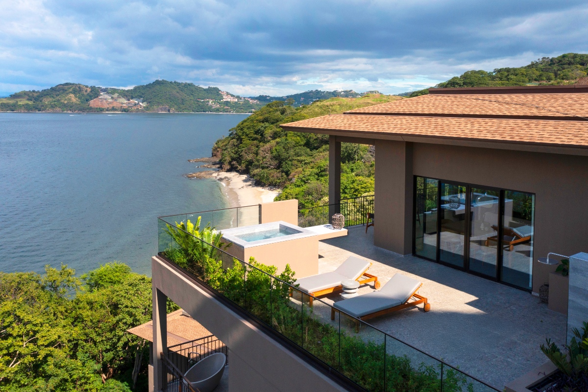 A private terrace with two wooden lounge chairs and a raised plunge pool, edged by a glass railing and backed by sliding glass doors that open into a suite with a tile-roofed overhang. Beyond the terrace, forested cliffs drop to a small sand beach and calm ocean water, with green hills and scattered hillside houses along the coastline under a cloudy sky.
