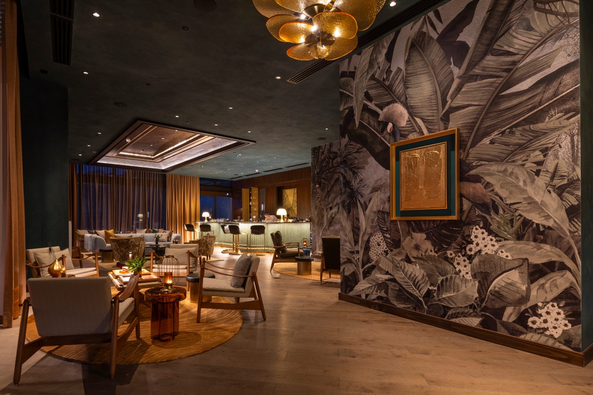 A dimly lit hotel lounge with clusters of wood-framed armchairs and low tables on round rugs, leading back to a bar with upholstered stools and a curtained wall of windows. A large-scale mural of tropical leaves and birds in muted gray tones covers the wall at right, hung with a gold framed artwork, and a brass petal-shaped chandelier hangs overhead.