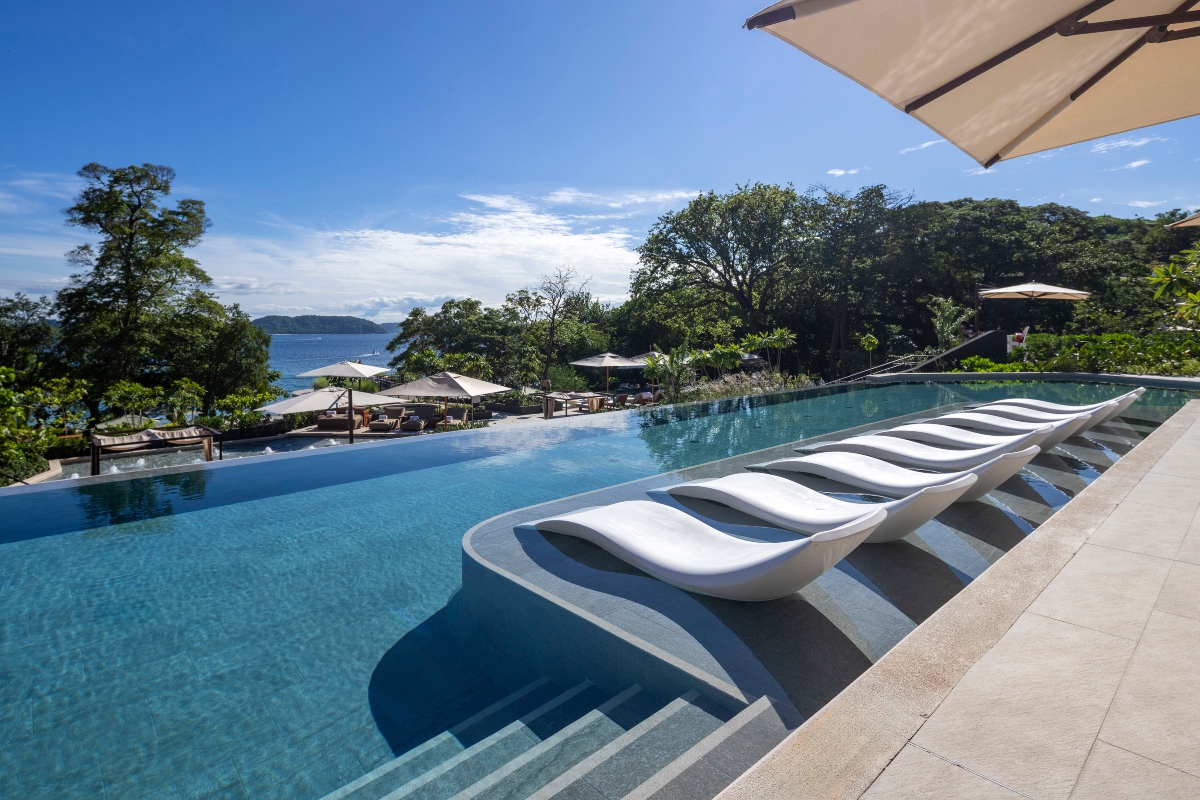 A resort infinity pool with a row of curved white in-water loungers set along a shallow ledge, steps descending into the water at center and a cream umbrella overhead at right. Beyond the pool's edge, shaded daybeds and more umbrellas line a terrace framed by tall green trees, with the ocean and a distant headland visible under a bright blue sky.