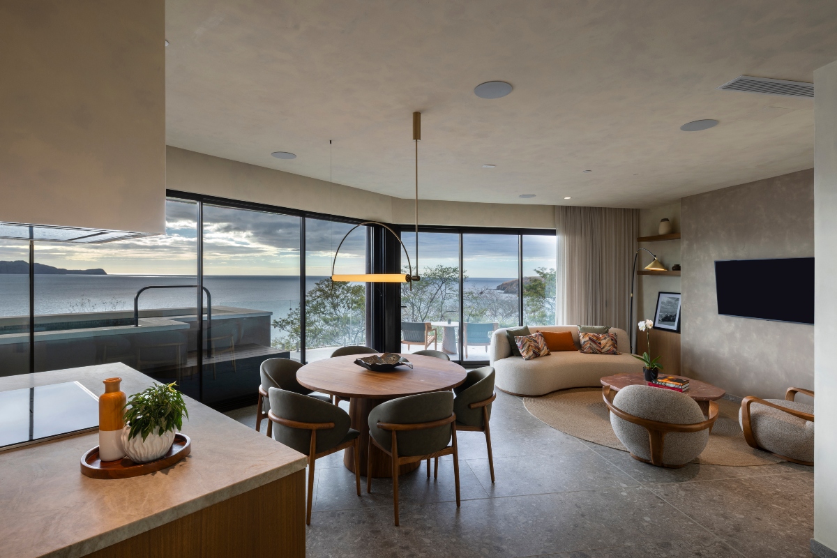 An open-plan residence interior with a stone-topped kitchen island in the foreground, a round wood dining table with six upholstered chairs beneath an arced pendant light, and a curved sofa with patterned pillows facing a wall-mounted television. Floor-to-ceiling glass doors wrap two walls and open onto a terrace with a pool and lounge chairs, framing a view of the ocean and a distant headland at dusk.