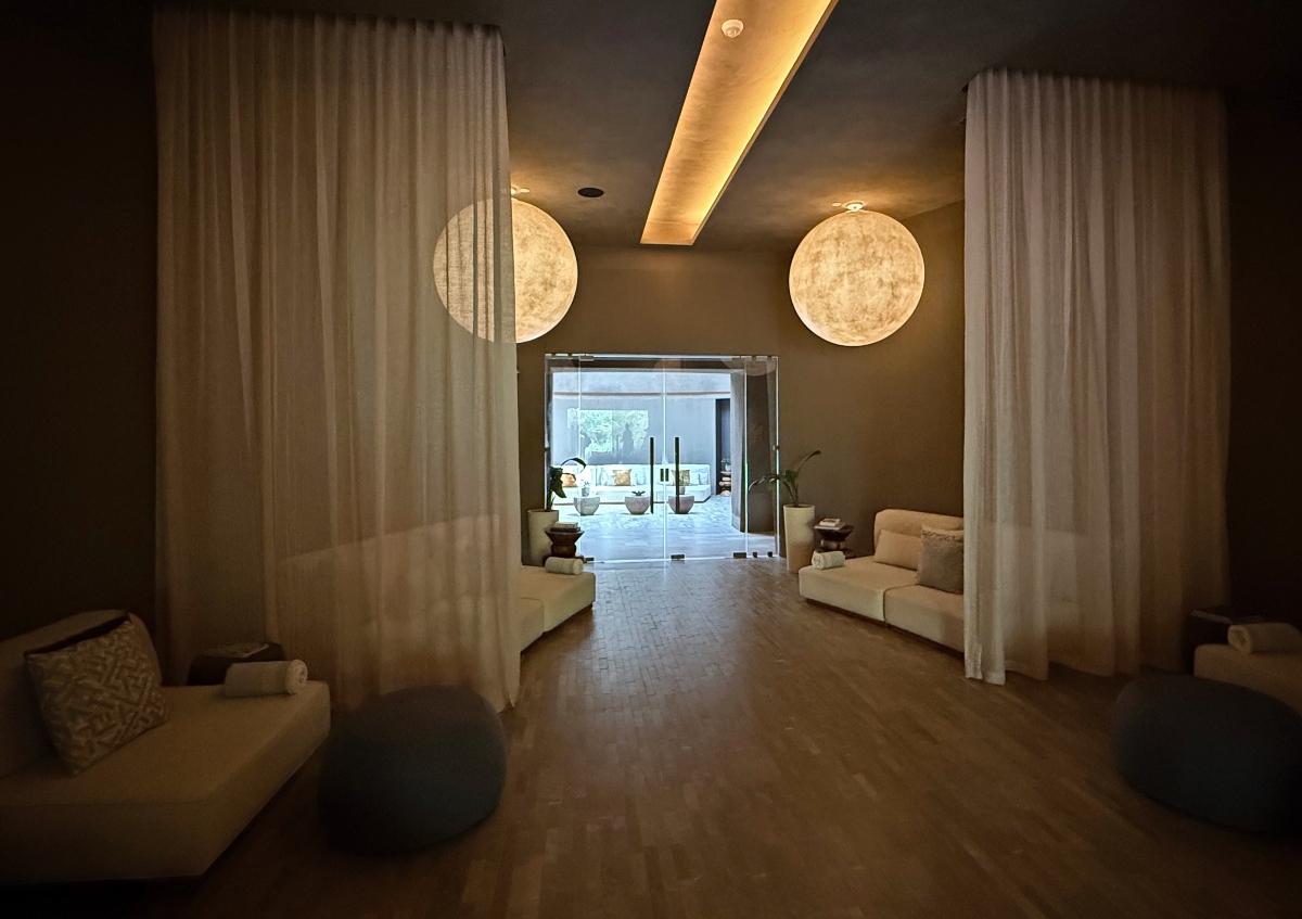 A dim spa relaxation lounge with low cream daybeds and round gray ottomans lining both sides of a wood floor, sheer curtains hanging from ceiling to floor between them. Two large round wall lights glow at either side of the room, and glass doors at the far end open onto a brighter seating area with potted plants.
