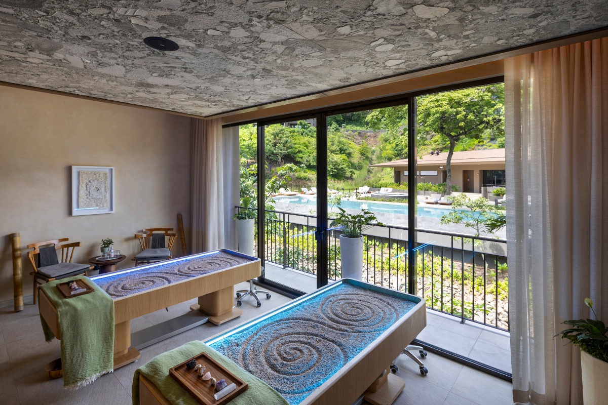 A spa treatment room with two side-by-side tables holding sand beds raked into spiral patterns, each lit from within and set with a green throw and a wood tray of crystals. Floor-to-ceiling windows and a sheer curtain open to a balcony view of a pool lined with loungers, a tile-roofed building, and green hillside vegetation, beneath a textured stone-patterned ceiling.
