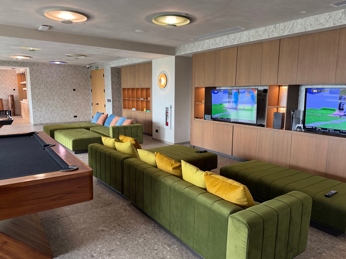A teen lounge with long channel-tufted olive green sofas and matching benches arranged in rows, scattered with yellow, blue, and pink pillows. Two large televisions displaying a video game are recessed into a wood cabinet wall on the right, and a pool table sits in the foreground at left.