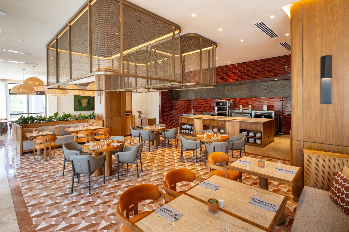 A restaurant dining room with wood tables set for service, surrounded by orange velvet and gray leather chairs on a patterned tile floor of terracotta and cream triangles. At the back, an open kitchen with a wood-fronted counter, stacked plates, and ovens sits against a red brick wall, while a large brass mesh light fixture hangs over the center of the room and woven pendant lamps light a banquette area to the left.