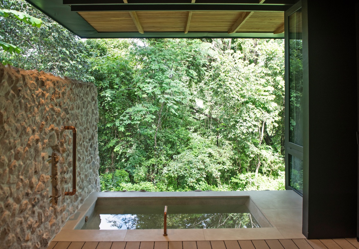 A private covered terrace with a rectangular sunken concrete plunge pool set into a wood-plank deck, its still water reflecting the surrounding trees. A rough stone wall with an exposed copper outdoor shower runs along the left side, and the open front of the terrace looks directly into dense green forest.