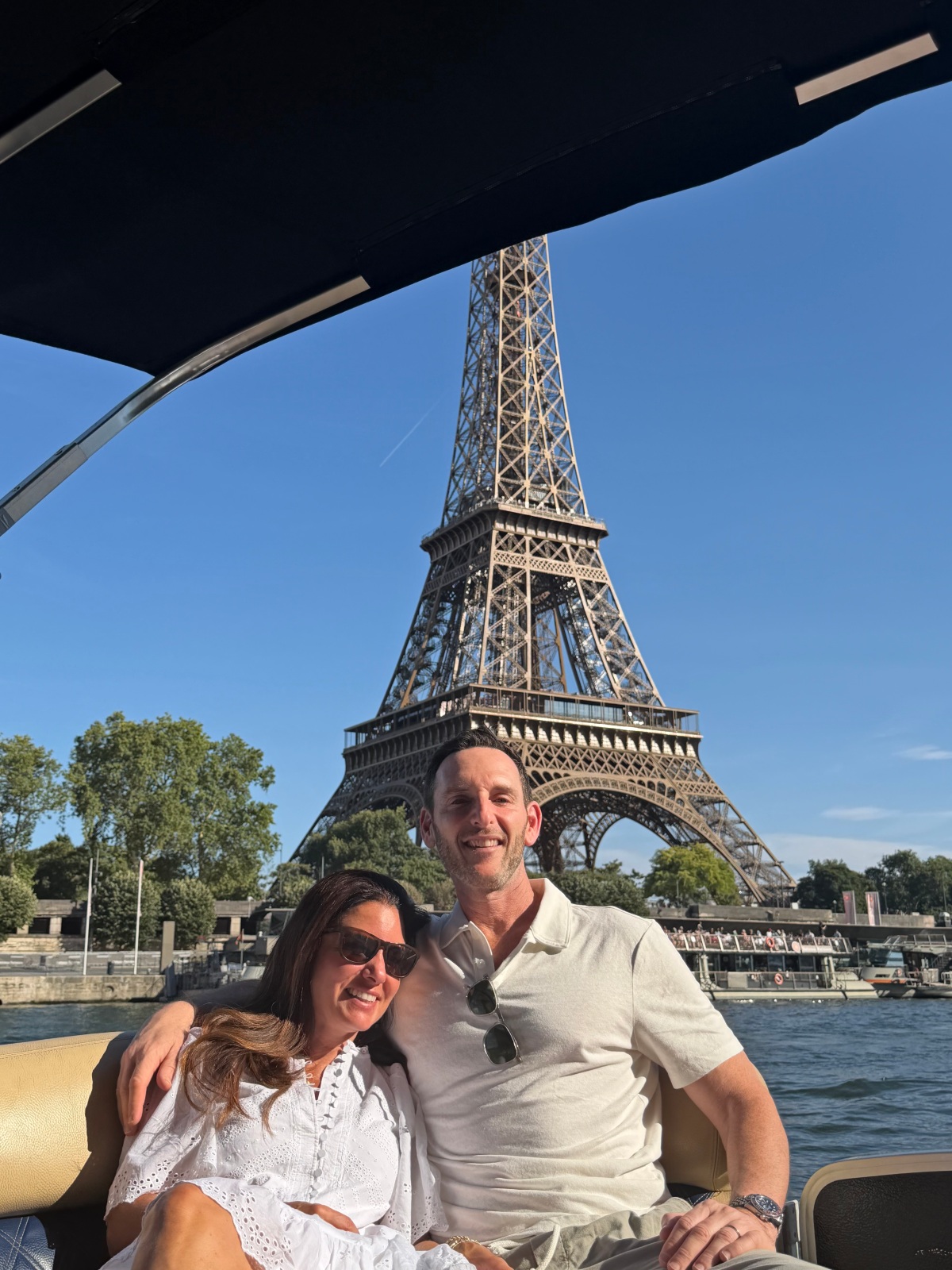 A smiling couple dressed in white sits close together on the cushioned seat of a small boat, the man's arm around the woman, who wears sunglasses and an eyelet dress. Behind them, the Eiffel Tower rises against a clear blue sky above the Seine River, with a tour boat docked along the tree-lined riverbank.