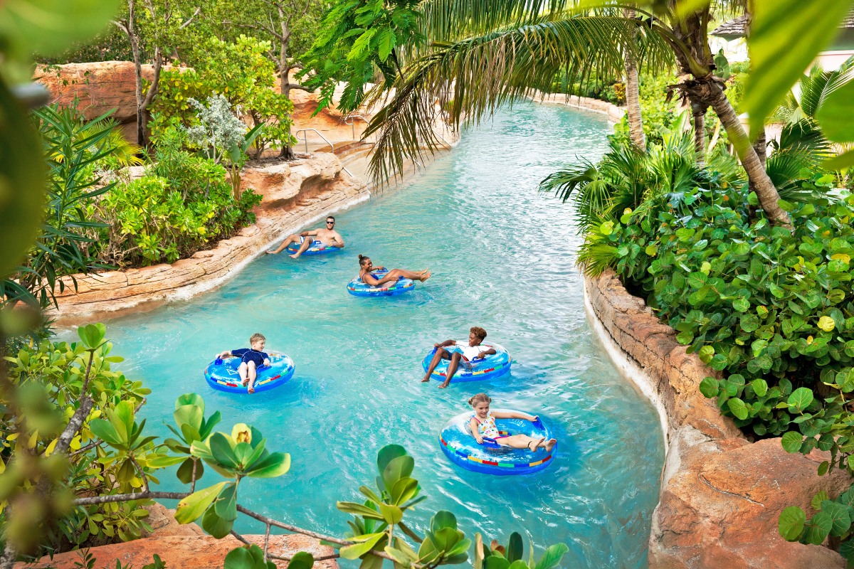 Five people, including two children, float on inflatable tubes along a winding lazy river bordered by palm trees, tropical plants, and rock formations. The water curves through lush landscaping under dappled sunlight, with everyone spaced along the current in individual tubes.