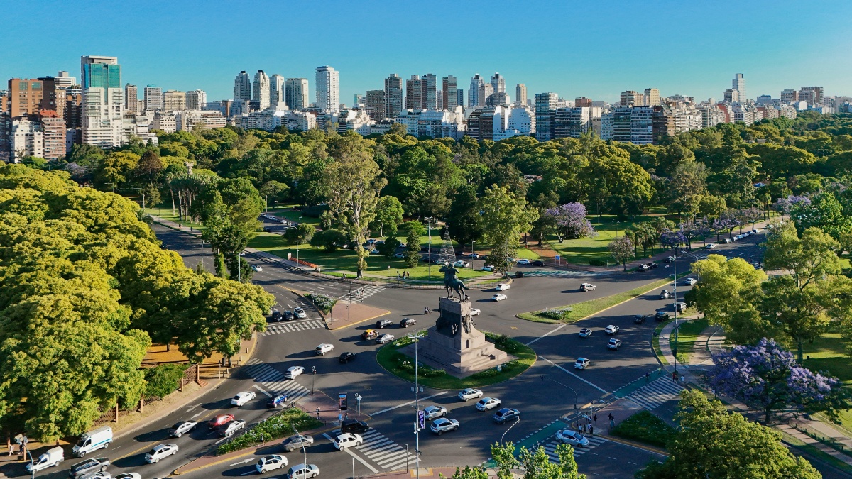 Aerial view of a large traffic roundabout with an equestrian monument at its center, surrounded by crosswalks and multiple lanes of car traffic. Green parkland with jacaranda trees in purple bloom stretches behind the roundabout, with a dense high-rise city skyline visible under a clear blue sky.