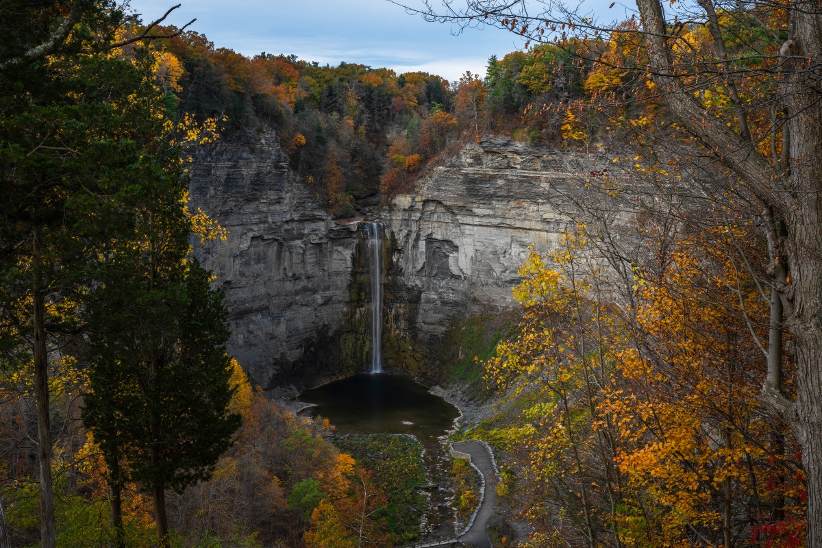 Taughannock Falls plunges over a tall, layered rock cliff into a pool at the bottom of a deep gorge in New York’s Finger Lakes region. Trees covered in orange, yellow and green autumn foliage frame the waterfall and rocky gorge.
