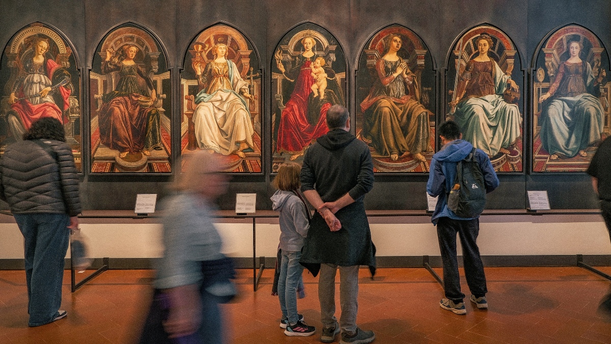 Museum visitors, some blurred in motion, stand and view a series of arched Renaissance panel paintings depicting a Madonna and Child alongside female saints or allegorical figures in ornate robes. The gold-toned religious artworks hang in a row along a dark wall, each with a small descriptive placard mounted below it.