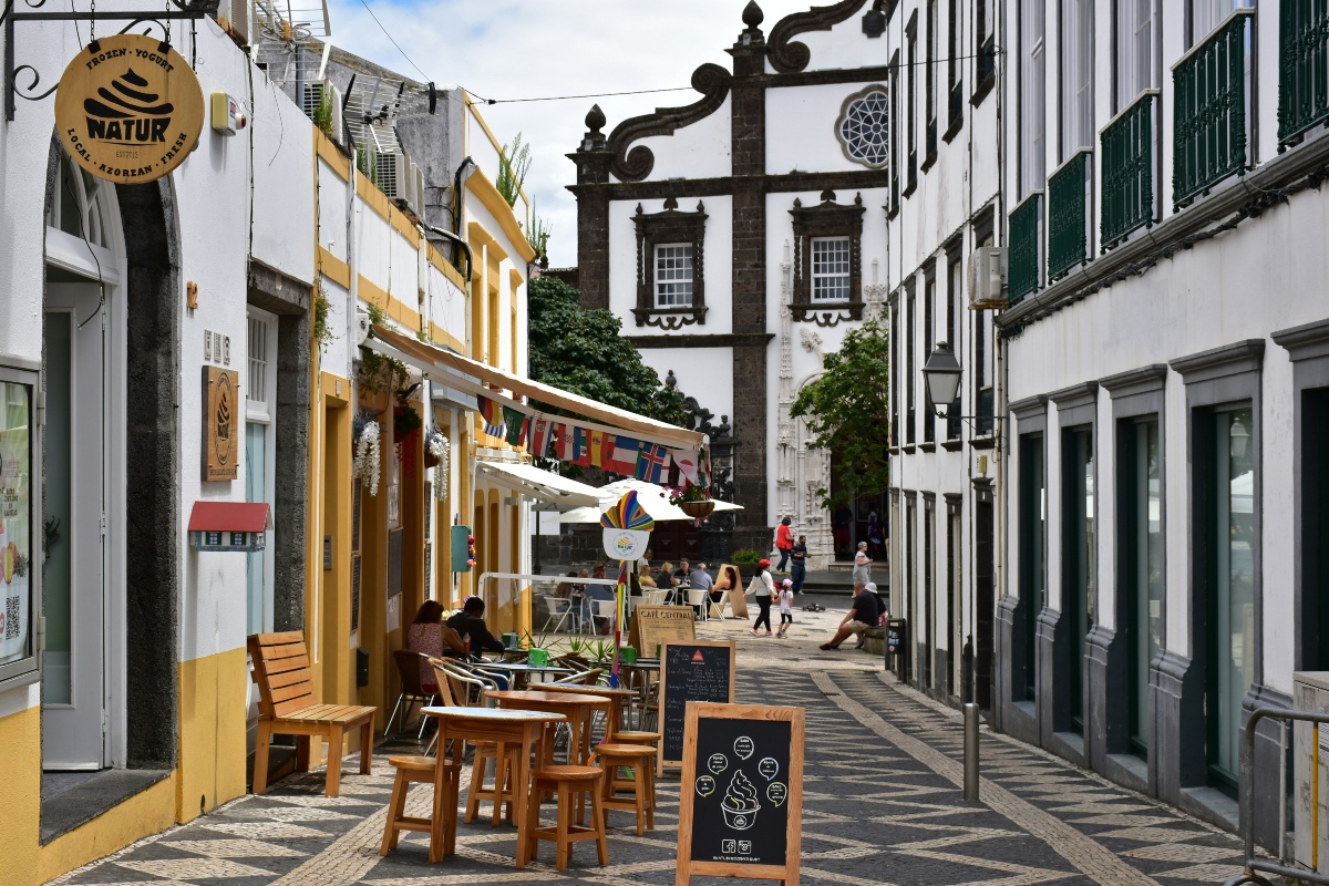 A narrow pedestrian street in Ponta Delgada is lined with cafés, outdoor seating and whitewashed buildings trimmed in dark volcanic stone. A black-and-white church façade rises at the end of the street, with people walking and sitting nearby.