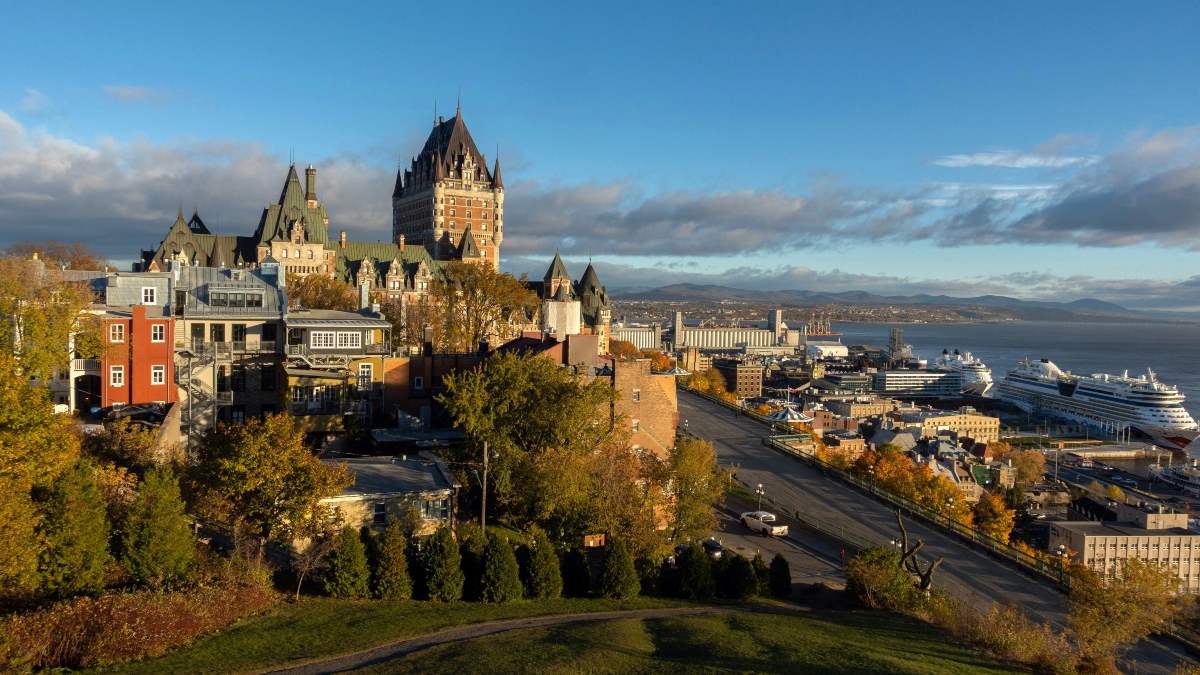 Aerial view of Château Frontenac's copper-green roofs and turrets rising above Quebec City's Old Town, with colorful row houses and autumn foliage in the foreground. The St. Lawrence River and two cruise ships are visible in the background under a partly cloudy blue sky.