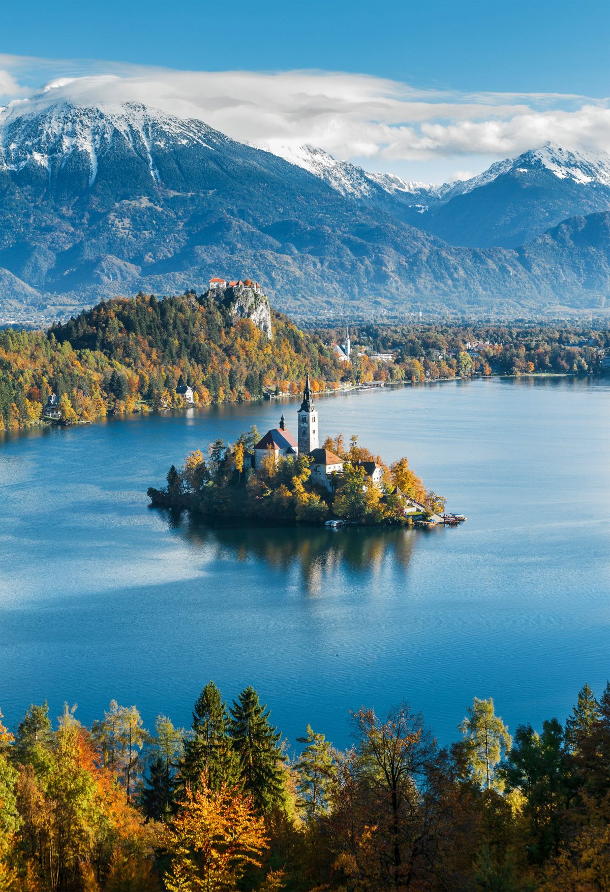 A small island with a church featuring a tall bell tower and red rooftops sits in the middle of a calm blue lake, surrounded by autumn-colored trees. Bled Castle perches on a cliff overlooking the lake, with snow-capped mountains rising in the background under a partly cloudy sky.