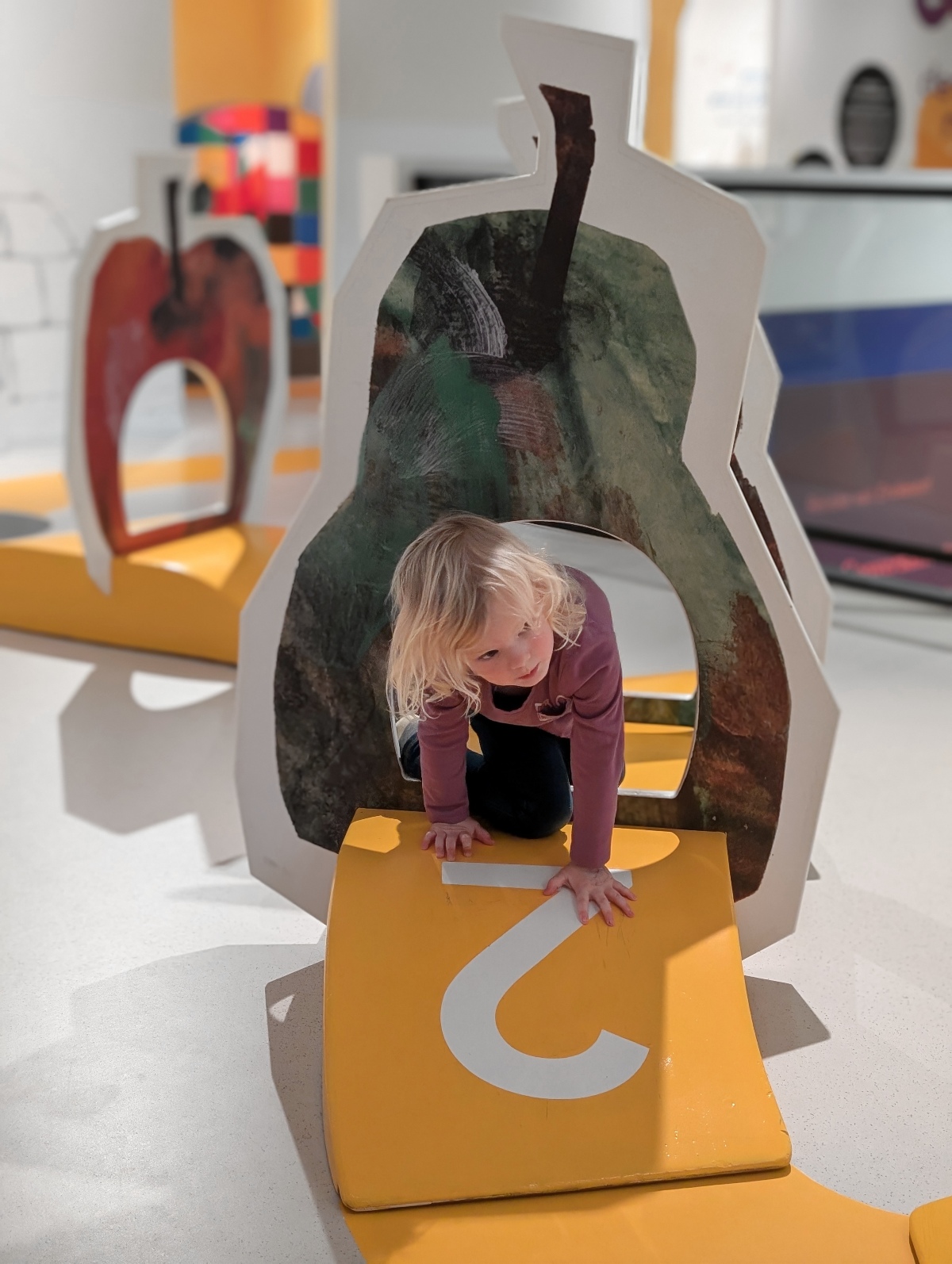 A young blond child in a long-sleeved purple shirt crawls through a round opening cut into a large painted cutout of a green and brown pear, placing their hands on a curved yellow ramp marked with a white number 2. Behind the child, a matching cutout of a red apple and a colorful patchwork-patterned panel stand in a bright indoor play space with white floors and yellow accents.