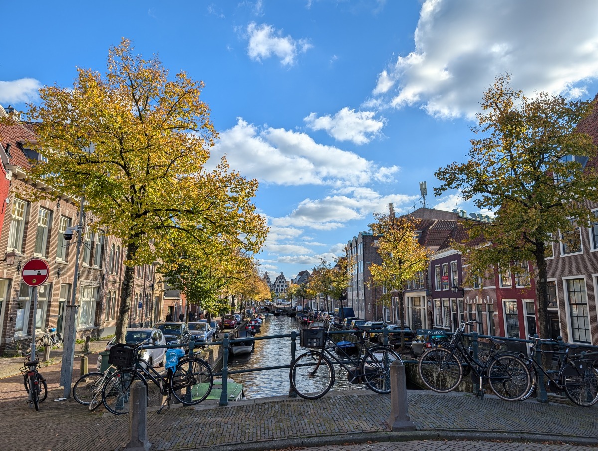 Several bicycles lean against a metal railing on a brick bridge overlooking a narrow canal lined with small boats, parked cars, and trees with yellow and green autumn leaves. Brick row houses with white-trimmed windows stretch along both sides of the canal toward a distant bridge and gabled buildings under a blue sky with puffy white clouds.