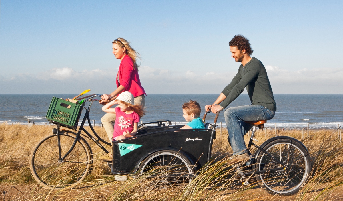 A family of four rides along a sandy coastal path lined with tall dune grass, with the sea and a clear blue sky in the background. A woman in a pink jacket pedals a bicycle with a green crate on the front, while a man in a dark green sweater pedals a cargo bike carrying a young girl in a white sun hat and a young boy in a teal shirt in its front box.