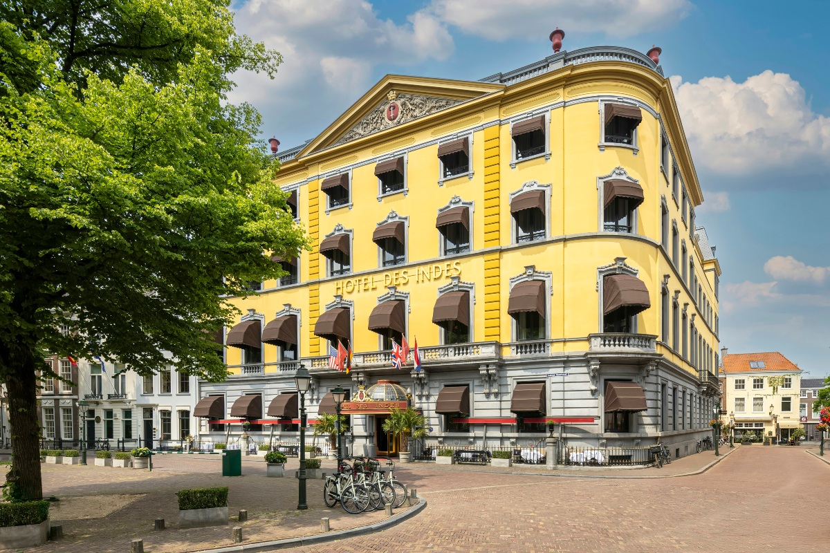 The yellow four-story facade of Hotel des Indes features ornate white window trim, brown striped awnings, a triangular pediment with a carved crest, and a row of international flags above its glass-canopied entrance. The hotel sits on a corner of a brick-paved square with a leafy tree on the left, parked bicycles, planters, and neighboring townhouses under a partly cloudy blue sky.