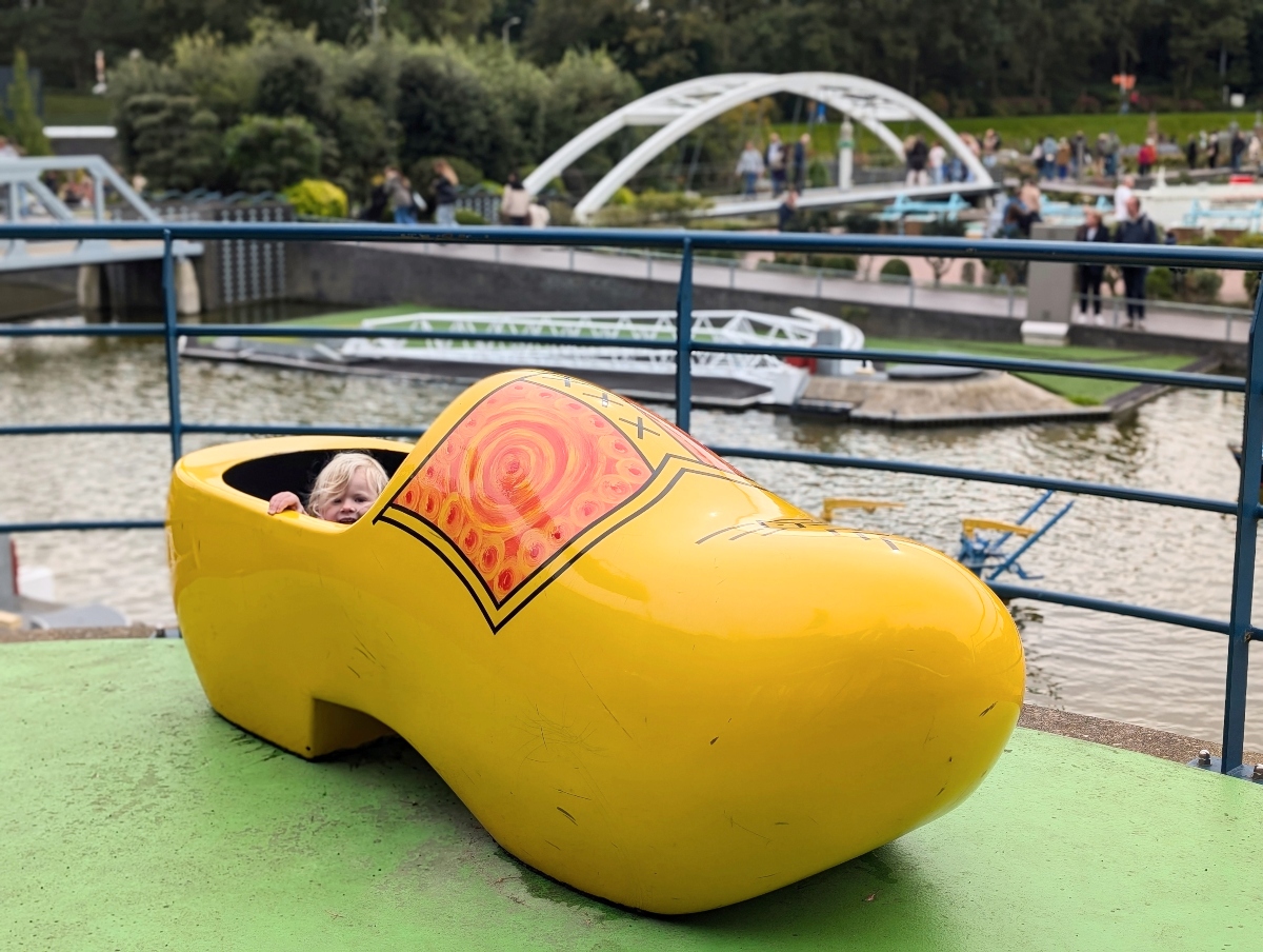 A young blond child peeks out from inside an oversized yellow wooden clog decorated with an orange swirl pattern, which sits on a green platform beside a blue metal railing. In the background, a canal holds miniature bridge and boat models, with a white arched bridge, trees, and visitors walking along the paths beyond.